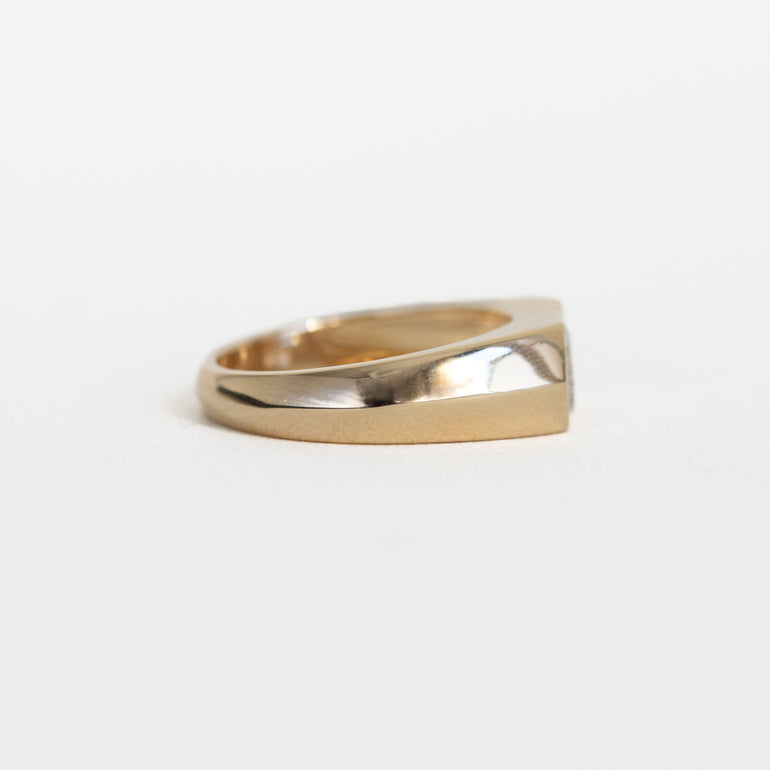 A gold signet-style ring with a wide, polished band and a flat, slightly angled face, photographed on a clean white background.