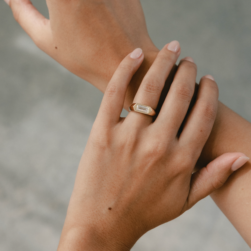 A close-up lifestyle shot of a person's hands, with one hand resting on the other and wearing a slim gold ring with a rectangular clear gemstone on the ring finger, set against a soft gray concrete background.