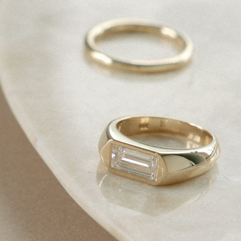 A polished yellow gold signet ring with a rectangular baguette-cut clear gemstone, displayed on a curved cream-colored stone surface next to a second plain gold ring.