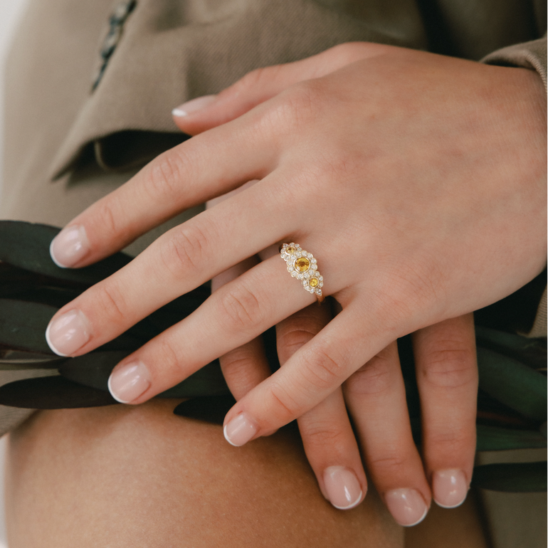 Hand wearing a gold ring with a floral design, holding black fabric against a neutral background.