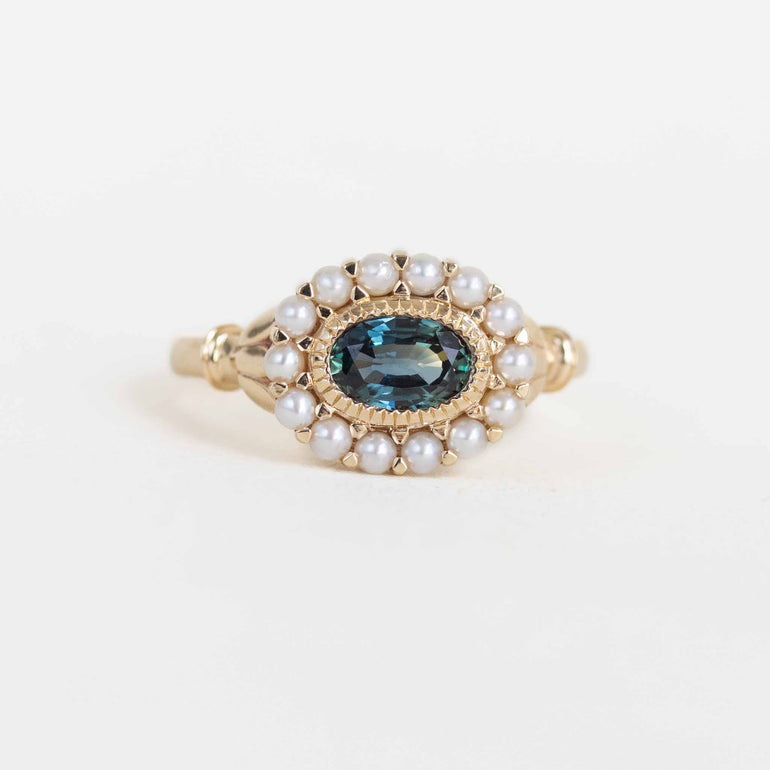 A gold ring featuring an oval blue gemstone surrounded by a halo of white pearls, set against a plain white background.