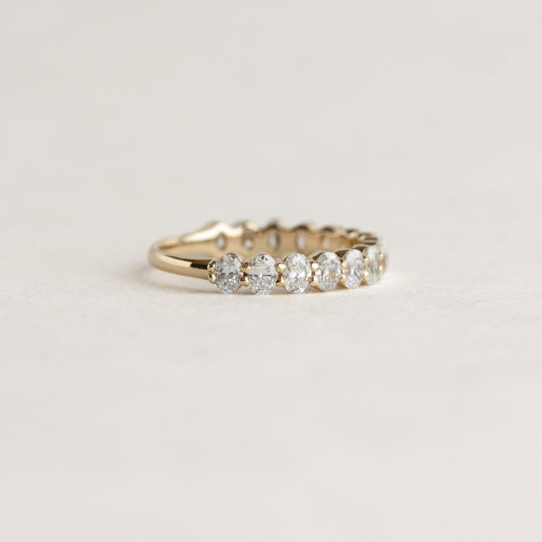 A yellow gold eternity ring featuring a row of round-cut clear gemstones, photographed on a clean, light beige textured background.