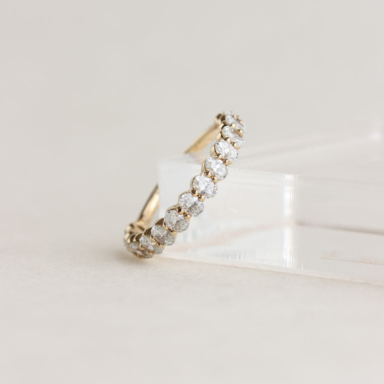 A yellow gold eternity ring set with round brilliant-cut diamonds, resting on a clear acrylic display block against a warm off-white background.