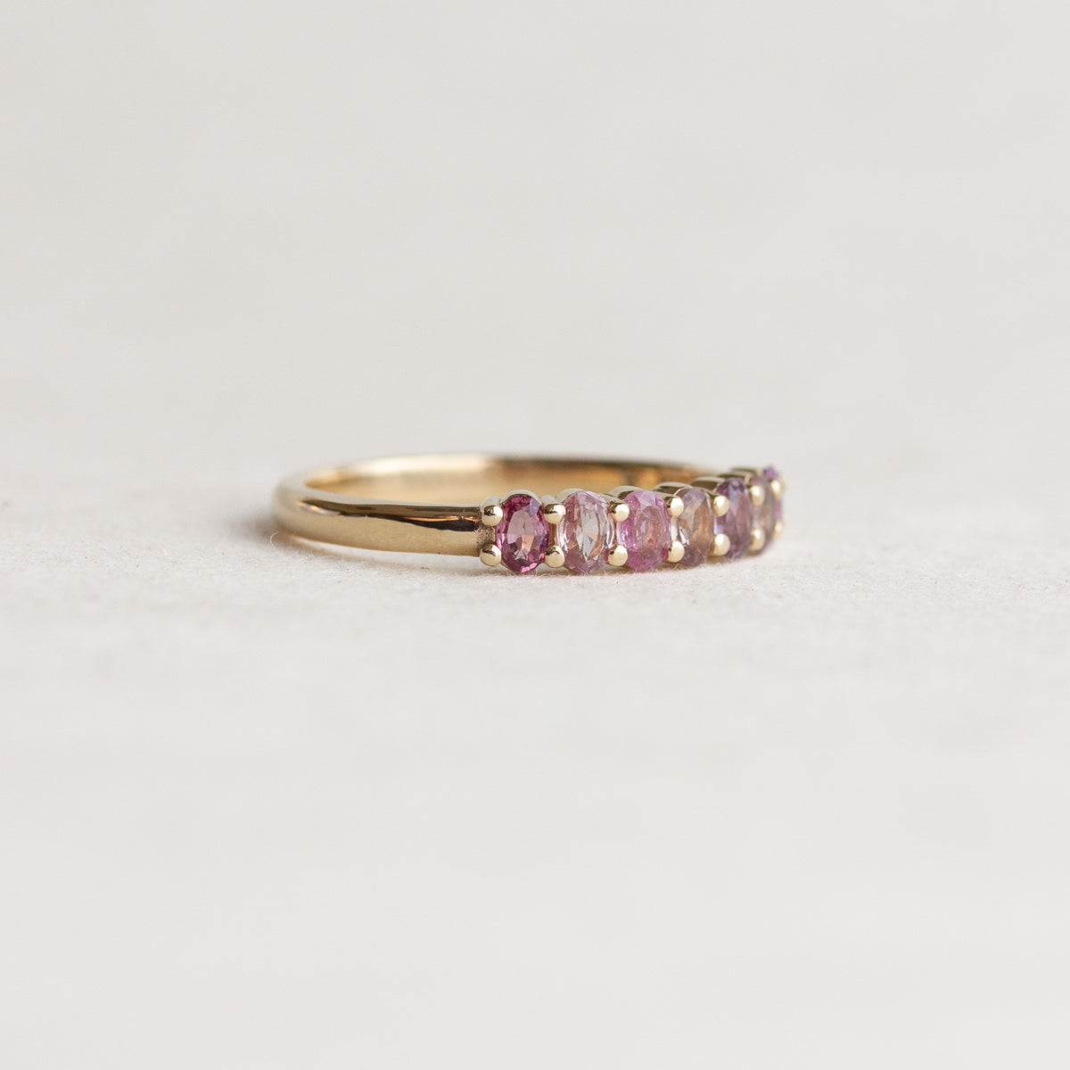 A gold band ring featuring a row of oval pink gemstones, photographed on a clean, light gray textured surface.