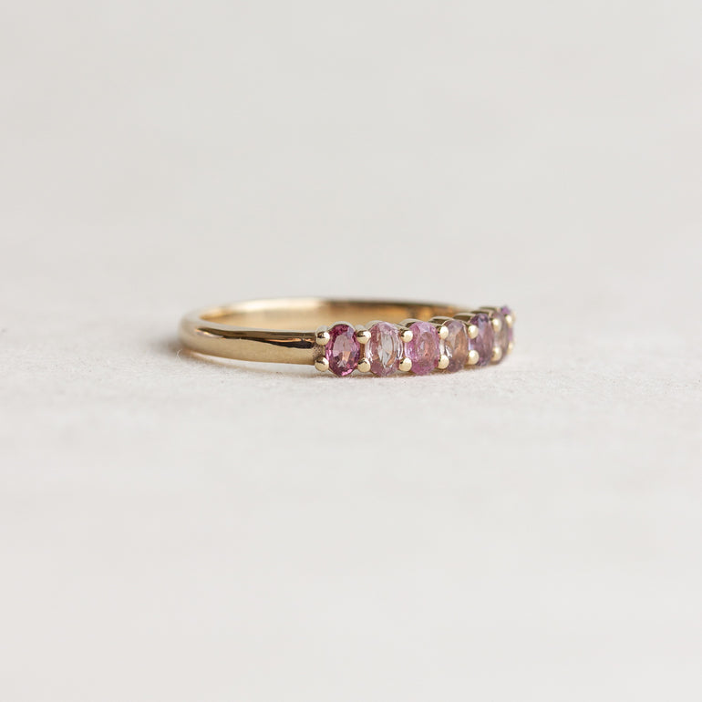 A gold band ring featuring a row of oval pink gemstones, photographed on a clean, light gray textured surface.