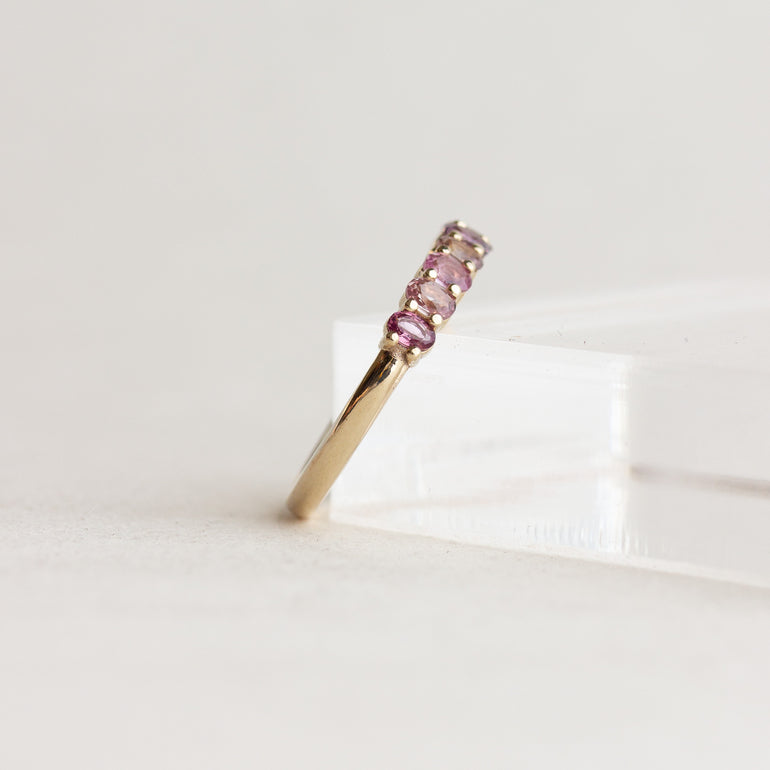 A gold ring with five small pink gemstones set in a row, displayed on a clear acrylic block against a light gray background.