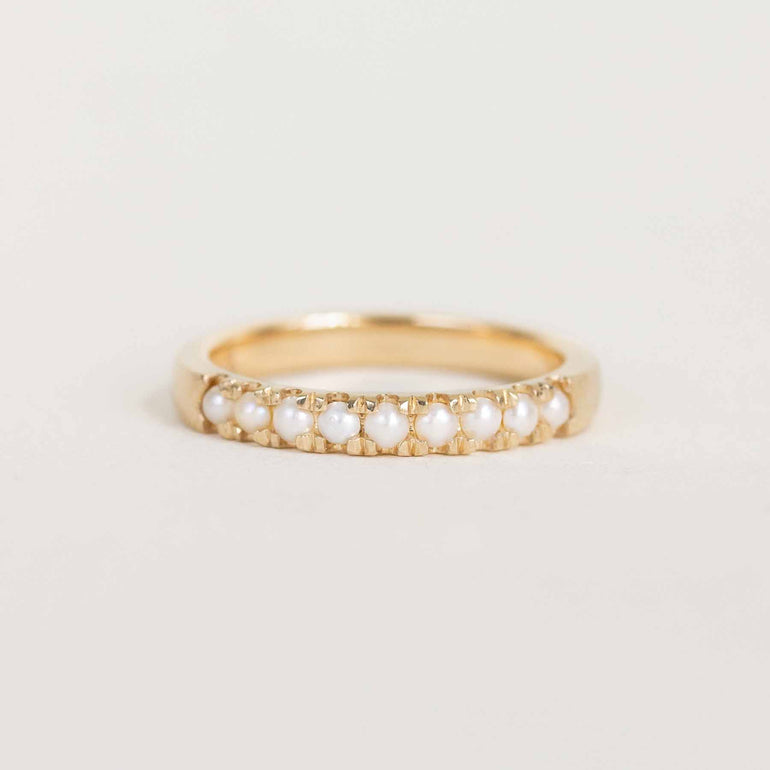A gold ring featuring a row of small white pearls set in a beaded gold border, photographed on a clean, light off-white background.