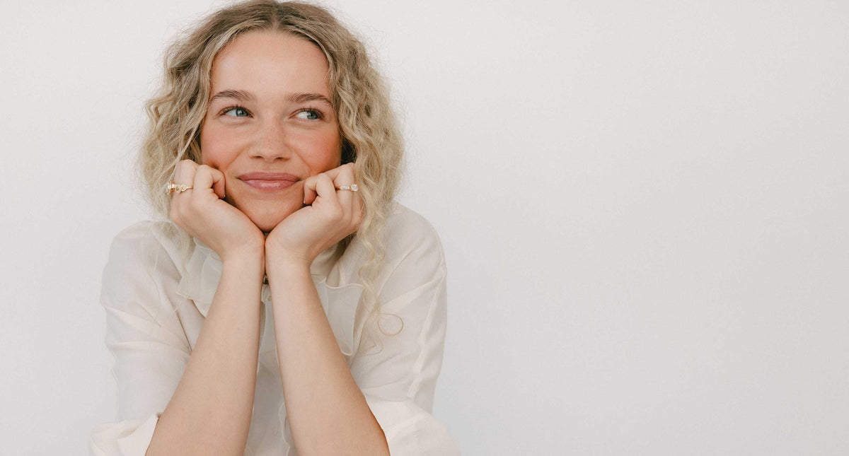A photo of a woman smiling with her hands under her chin, wearing multiple rings