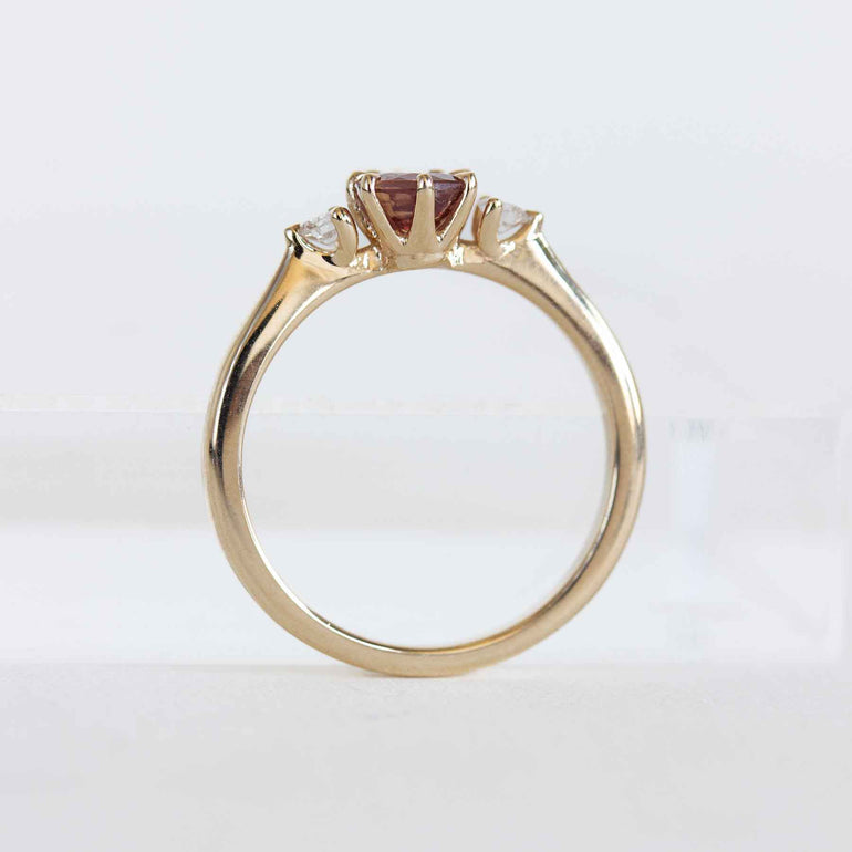 Gold ring with a berry sapphire and two smaller diamonds on a light gray background.