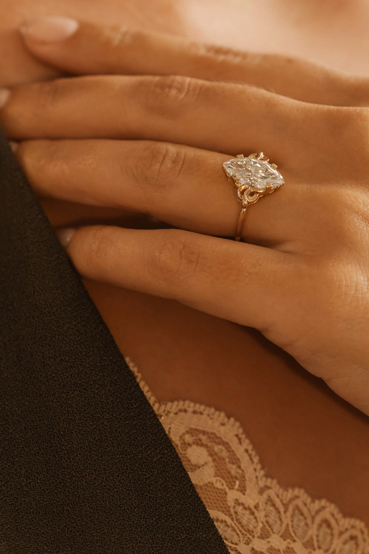 A close-up of a hand wearing a yellow gold ring with a large, multi-pronged clear gemstone, set against a warm, softly lit background with a lace garment visible at the bottom.