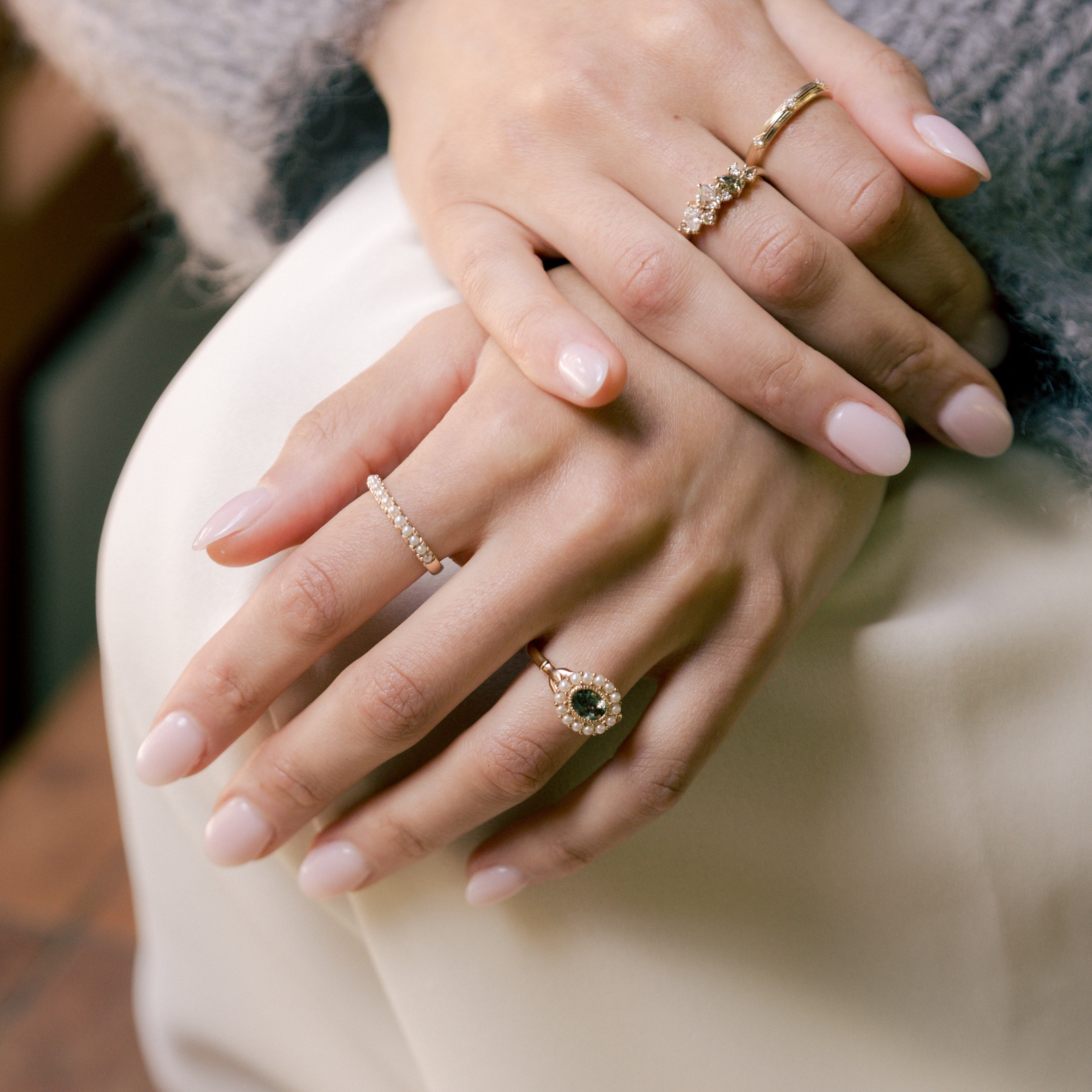 A close-up lifestyle shot of a person's hands resting on their lap, wearing several delicate gold rings, including a prominent ring with a dark green oval gemstone.