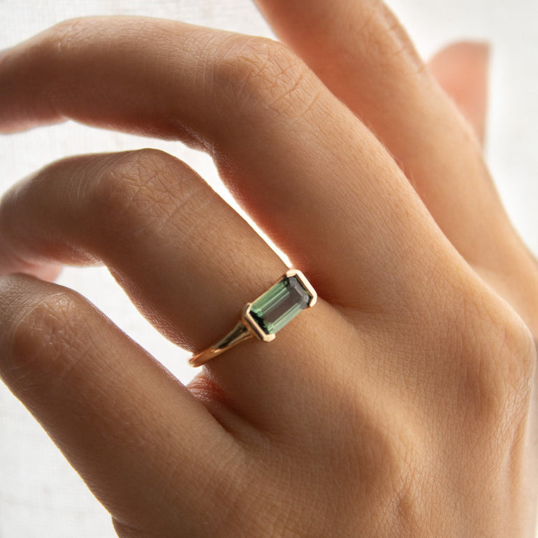 A close-up of a hand wearing a gold ring with a rectangular green gemstone, set against a soft, light-colored textured background.