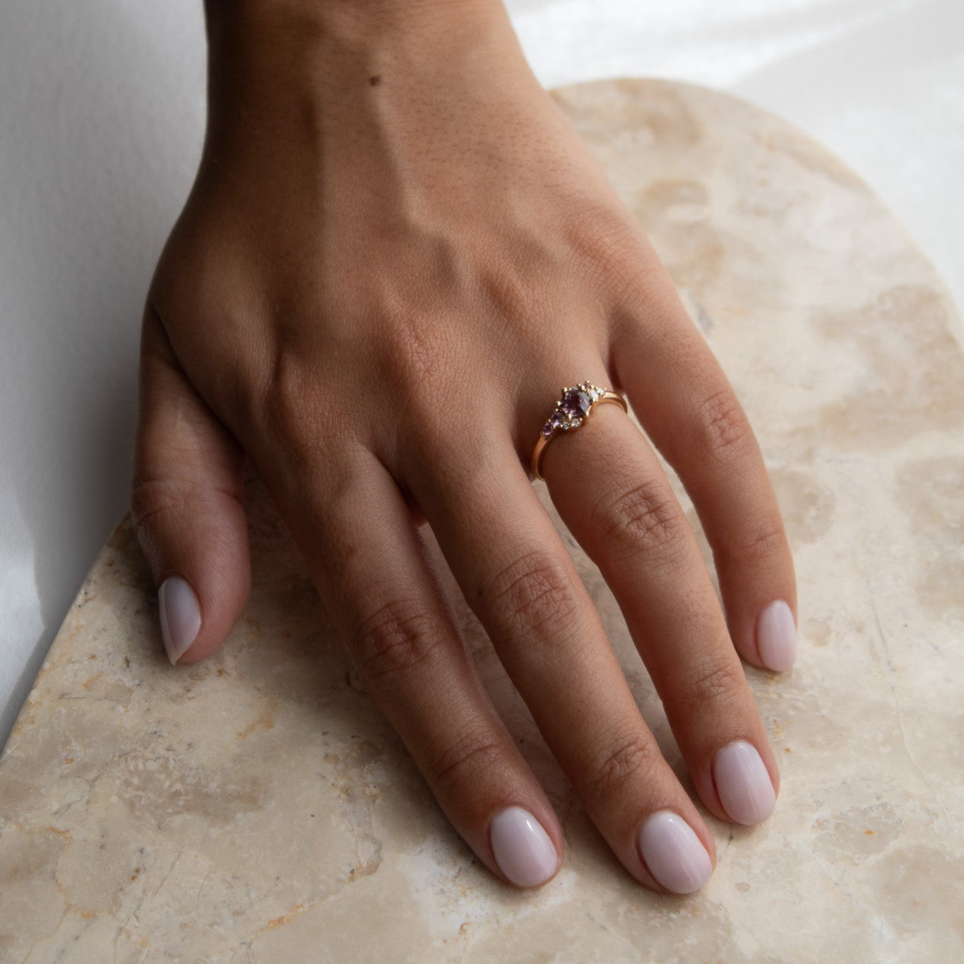 A close-up of a hand wearing a delicate gold ring with a marquise-cut purple gemstone, resting on a textured beige surface.