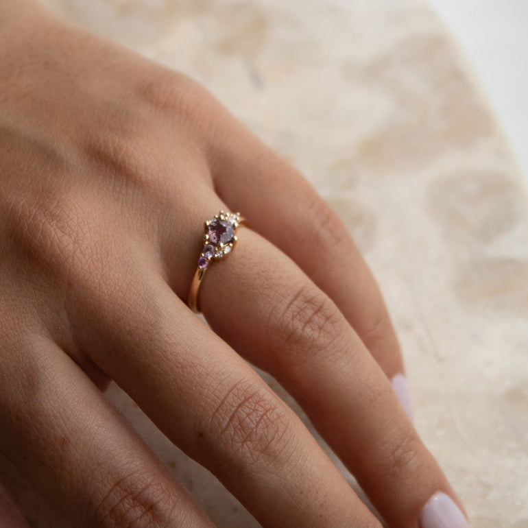 A close-up of a hand wearing a delicate gold ring with a marquise-cut purple gemstone, resting on a textured beige surface.