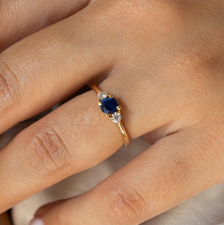 A close-up of a hand wearing a delicate gold ring with a deep blue oval center stone and small clear accent stones, set against a soft light fabric background.