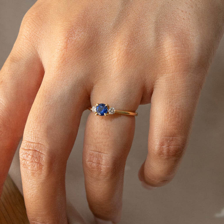 A close-up of a person's hand wearing a gold ring with a round blue gemstone and a small clear accent stone, resting on a light fabric surface.