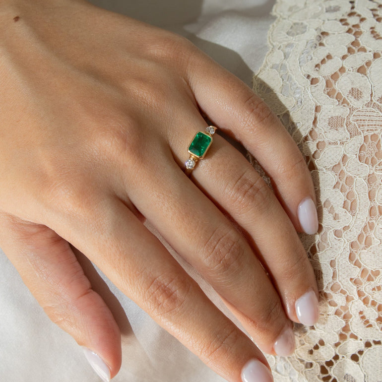 A close-up of a hand wearing a gold ring with a rectangular green gemstone, resting against white fabric and lace.