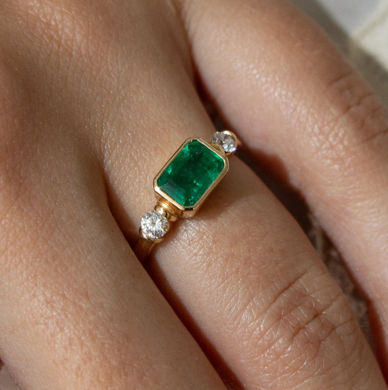 A close-up of a hand wearing a gold ring with a rectangular green gemstone, resting against white fabric and lace.