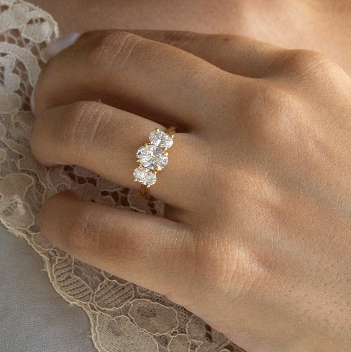 A close-up of a hand wearing a yellow gold ring with three clear marquise-cut gemstones, resting on a white lace-trimmed garment.