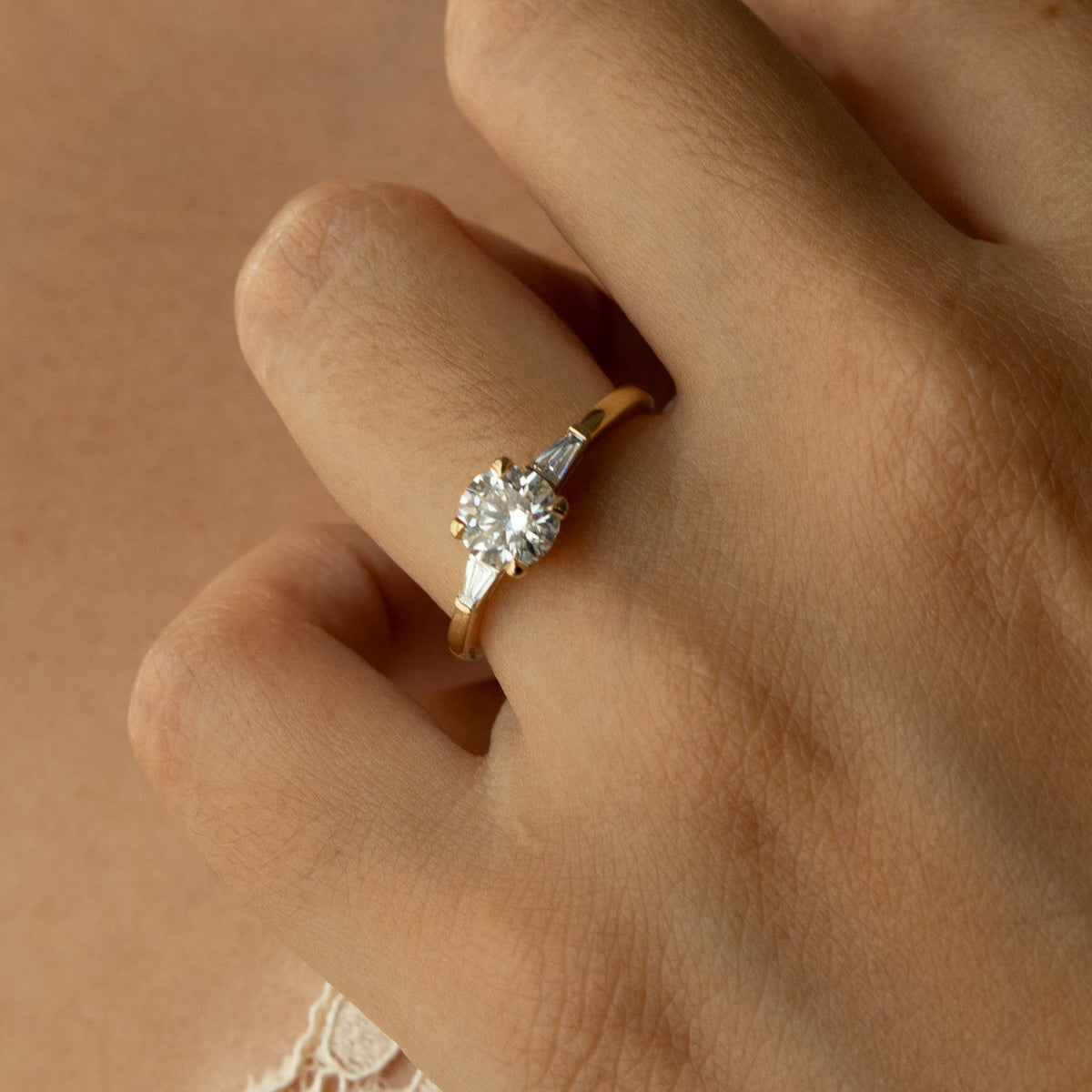 A close-up of a hand wearing a yellow gold solitaire engagement ring with a round-cut clear gemstone, set against warm skin tones and a hint of lace fabric.