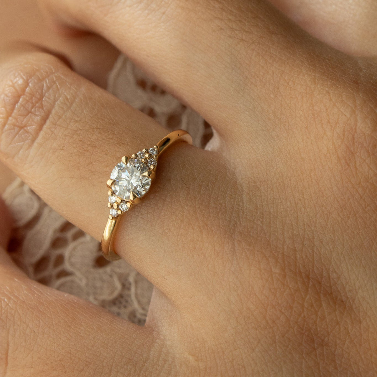 A close-up of a hand wearing a gold ring with a round clear center stone and smaller side stones, resting on a light lace fabric.