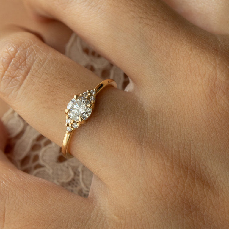 A close-up of a hand wearing a gold ring with a round clear center stone and smaller side stones, resting on a light lace fabric.