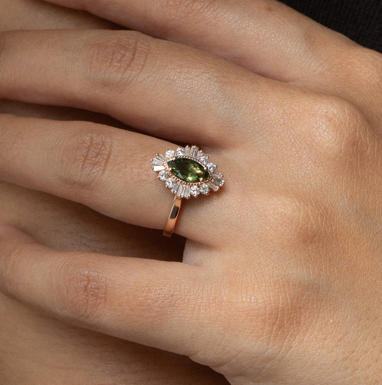 A close-up of a hand wearing a rose gold ring with a marquise-cut green gemstone surrounded by small clear stones, resting on the forearm.