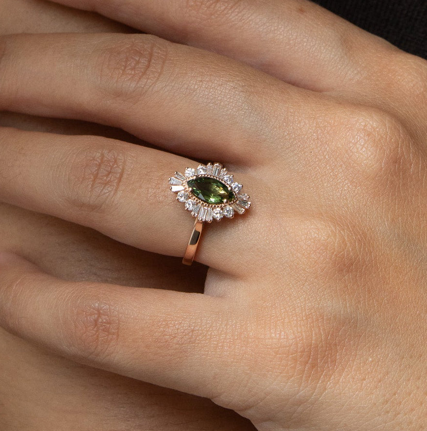 A close-up of a hand wearing a rose gold ring with a marquise-cut green gemstone surrounded by small clear stones, resting on the forearm.