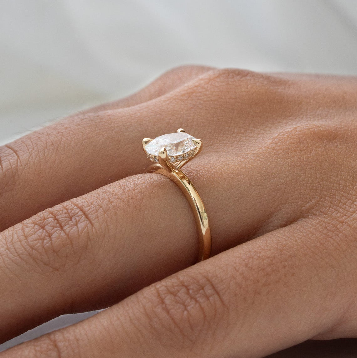 A gold ring with a marquise-cut clear gemstone worn on a person's finger, shown against a soft white draped fabric background.