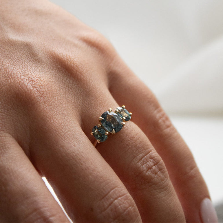 A close-up of a hand wearing a gold ring with three oval-cut blue gemstones, set against a soft, out-of-focus white background.