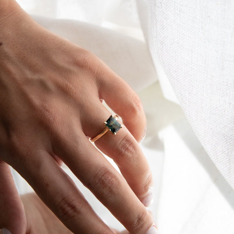 A close-up lifestyle shot of a person's hand wearing a gold ring with a dark green rectangular gemstone, set against a soft white fabric background.