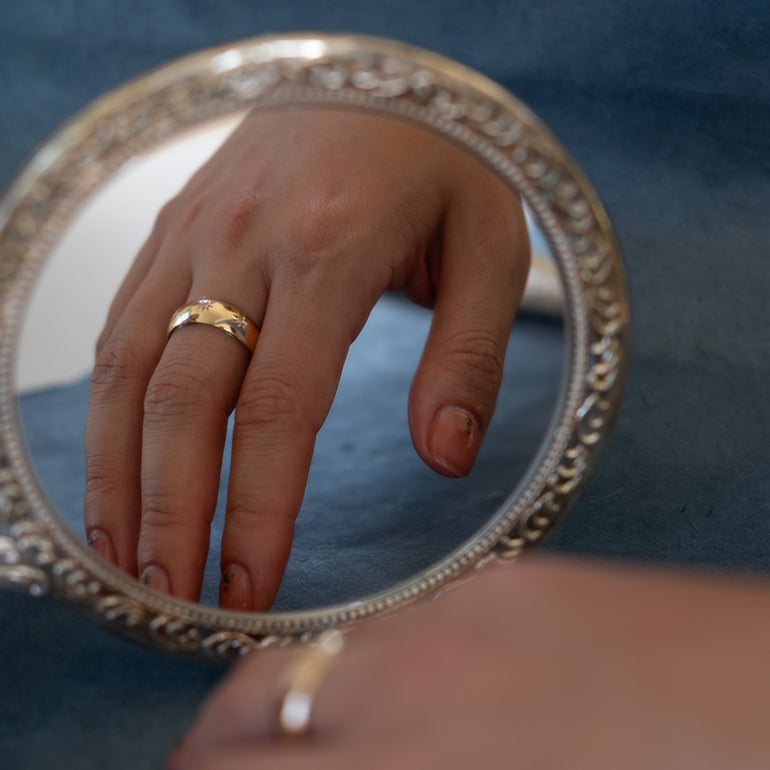 Hand with a gold ring held up to a decorative mirror against a blue background