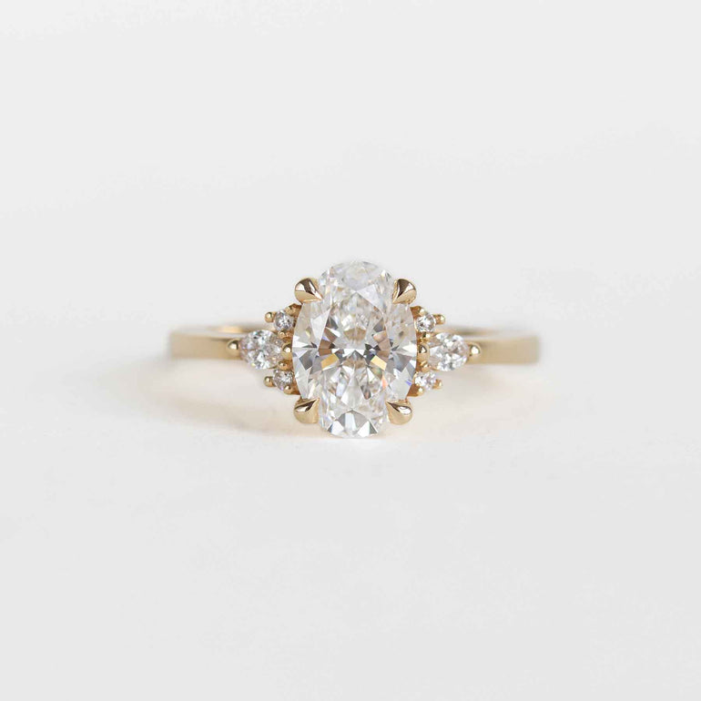 A yellow gold engagement ring featuring a large oval-cut center diamond surrounded by smaller round diamonds on the shoulders, set against a clean white background.