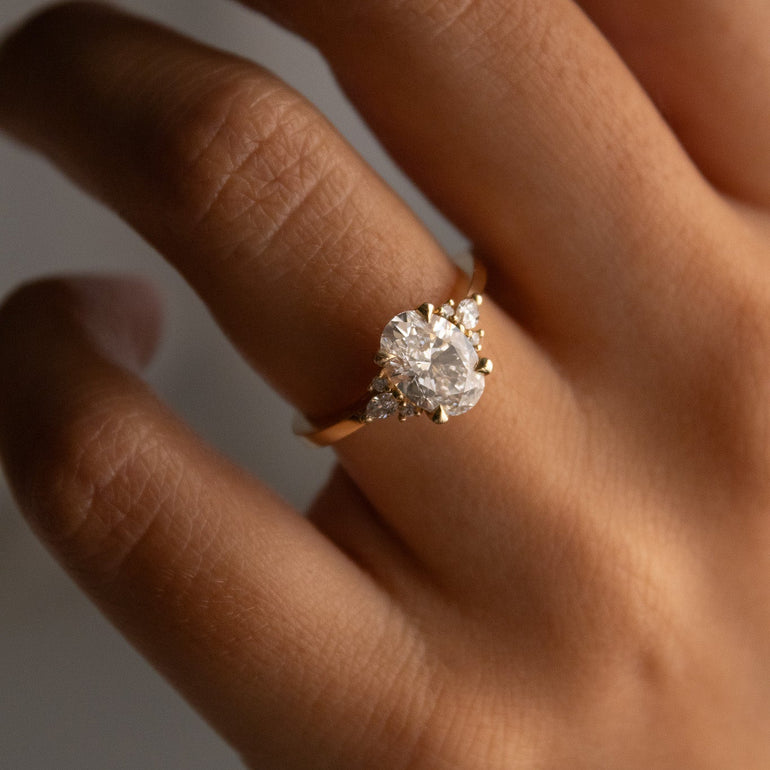 A close-up of a hand wearing a gold ring with a large lab diamond and smaller side diamonds, photographed against a softly blurred neutral background.