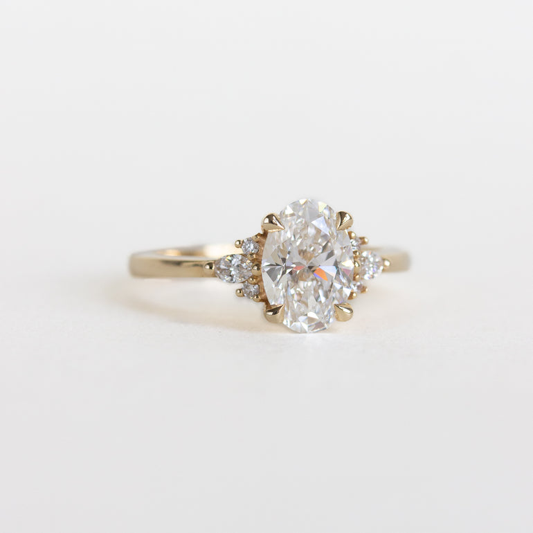 A yellow gold ring featuring a large oval-cut lab diamond with two smaller round-cut diamonds on either side of the band, set against a plain white background.