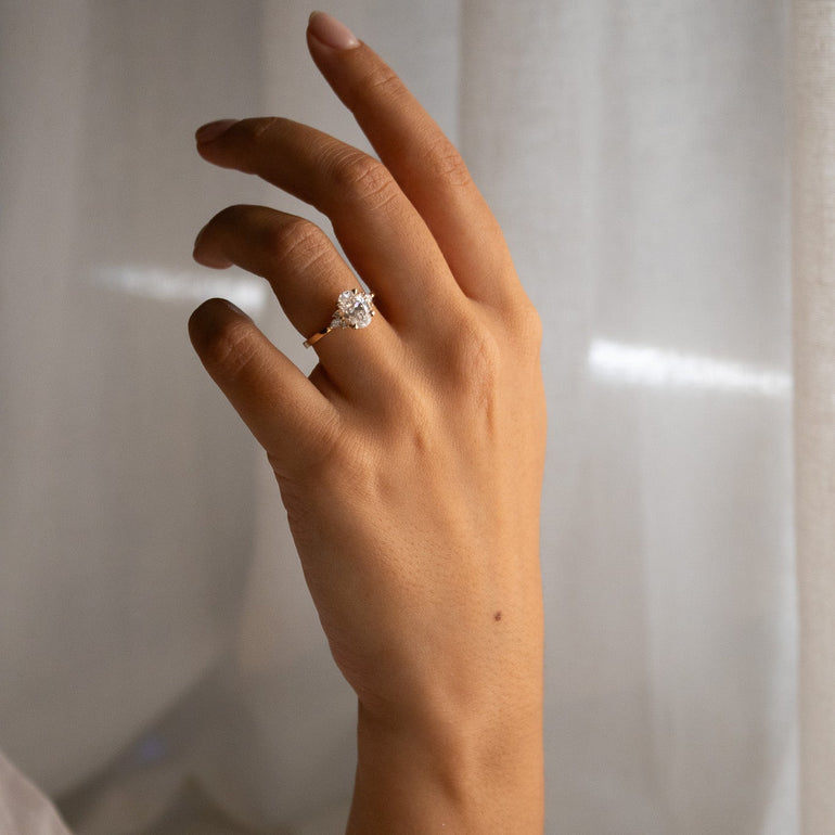 A close-up of a person's hand wearing a delicate gold ring with a diamond, set against a soft, white draped fabric background.