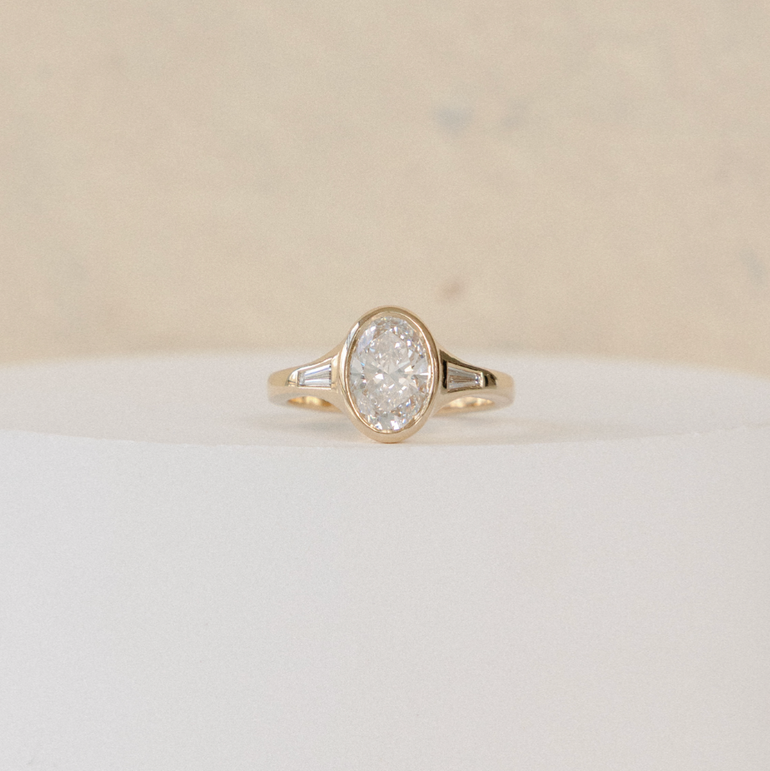 A gold oval ring with a clear round-cut center stone and small side stones, displayed on a white pedestal against a warm beige background.