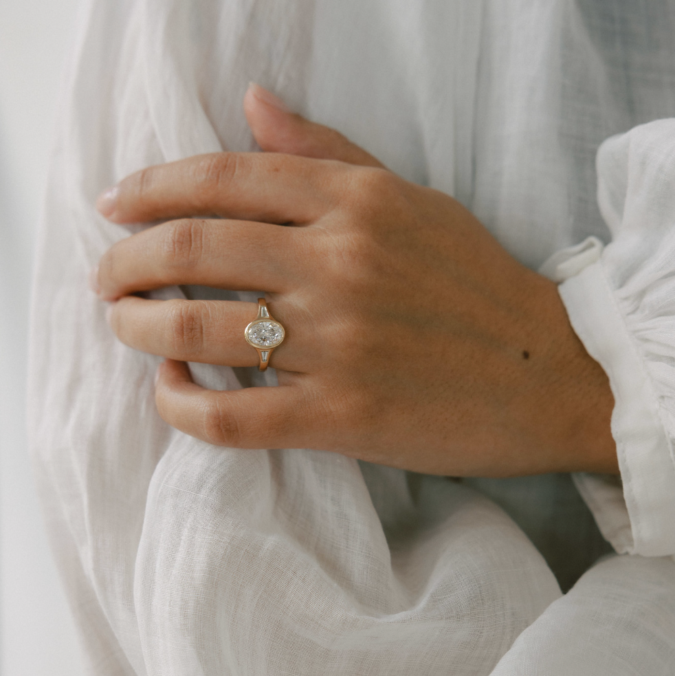 A close-up of a person's hand wearing a gold ring with a round clear gemstone, resting against white textured fabric.