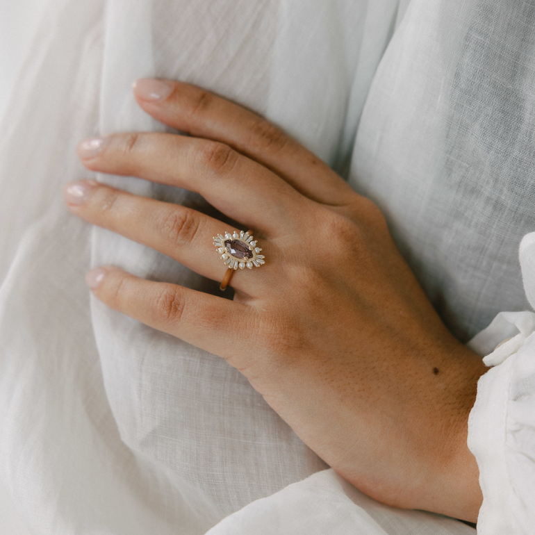 A close-up of a person's hand resting on white draped fabric, wearing a gold ring with a marquise-shaped purple amethyst center stone and small clear accent stones.