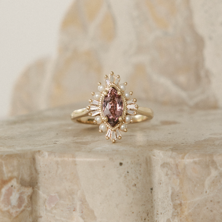 A gold ring with an oval pink gemstone and small clear accent stones, displayed on a beige stone surface with a softly blurred stone background.