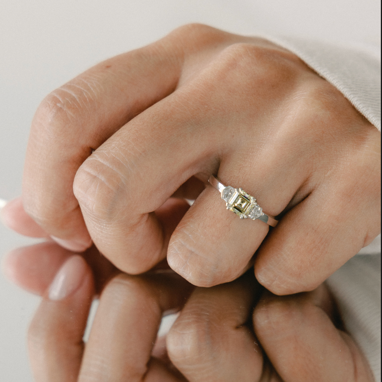 A close-up of a person's hands wearing a silver ring with a rectangular yellow gemstone and smaller clear accent stones, shown against a soft light background with a mirror reflection below.