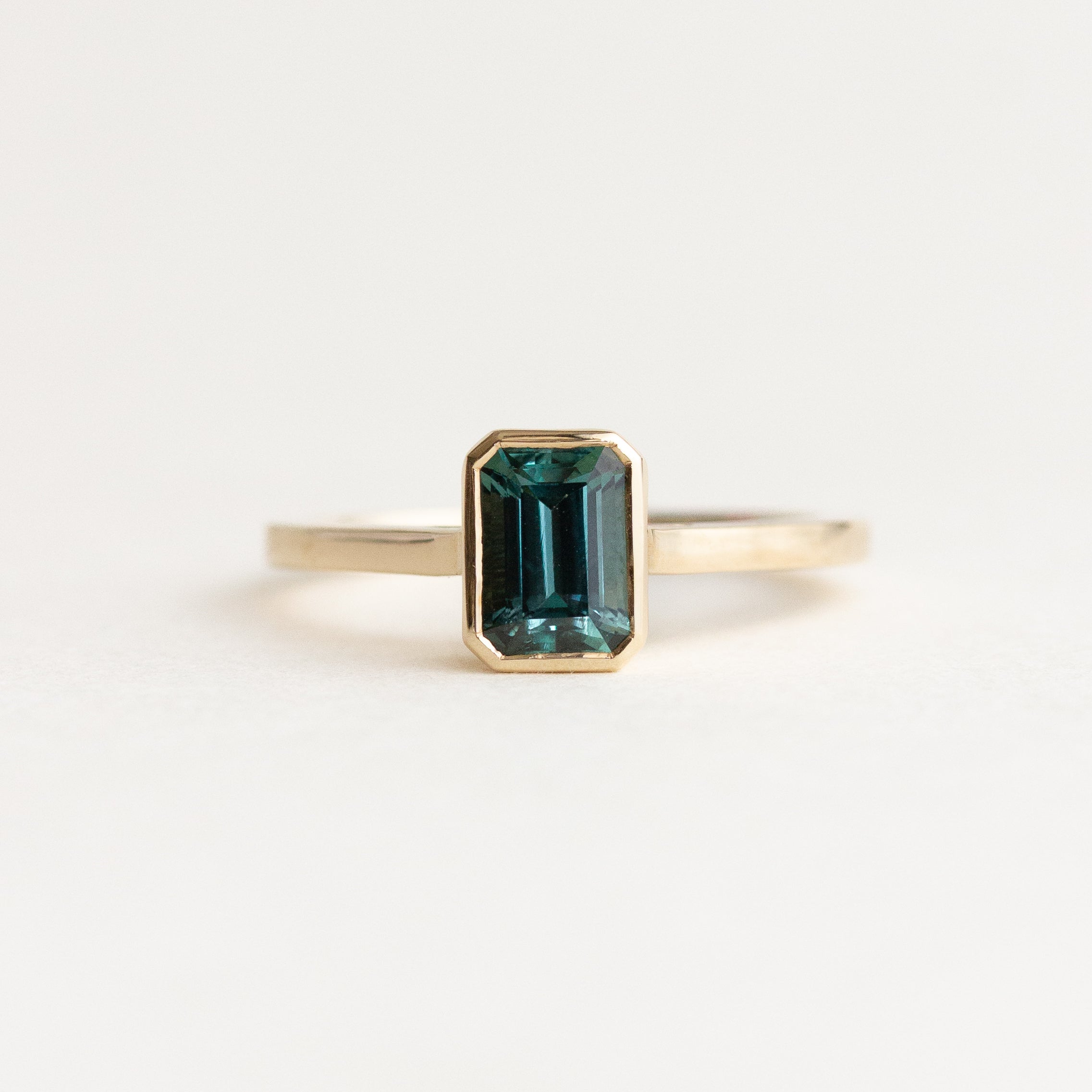 A gold ring featuring an emerald-cut dark teal green gemstone, centered on a clean white background.