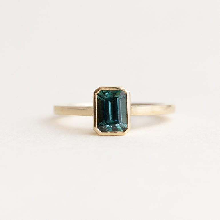 A gold ring featuring an emerald-cut dark teal green gemstone, centered on a clean white background.