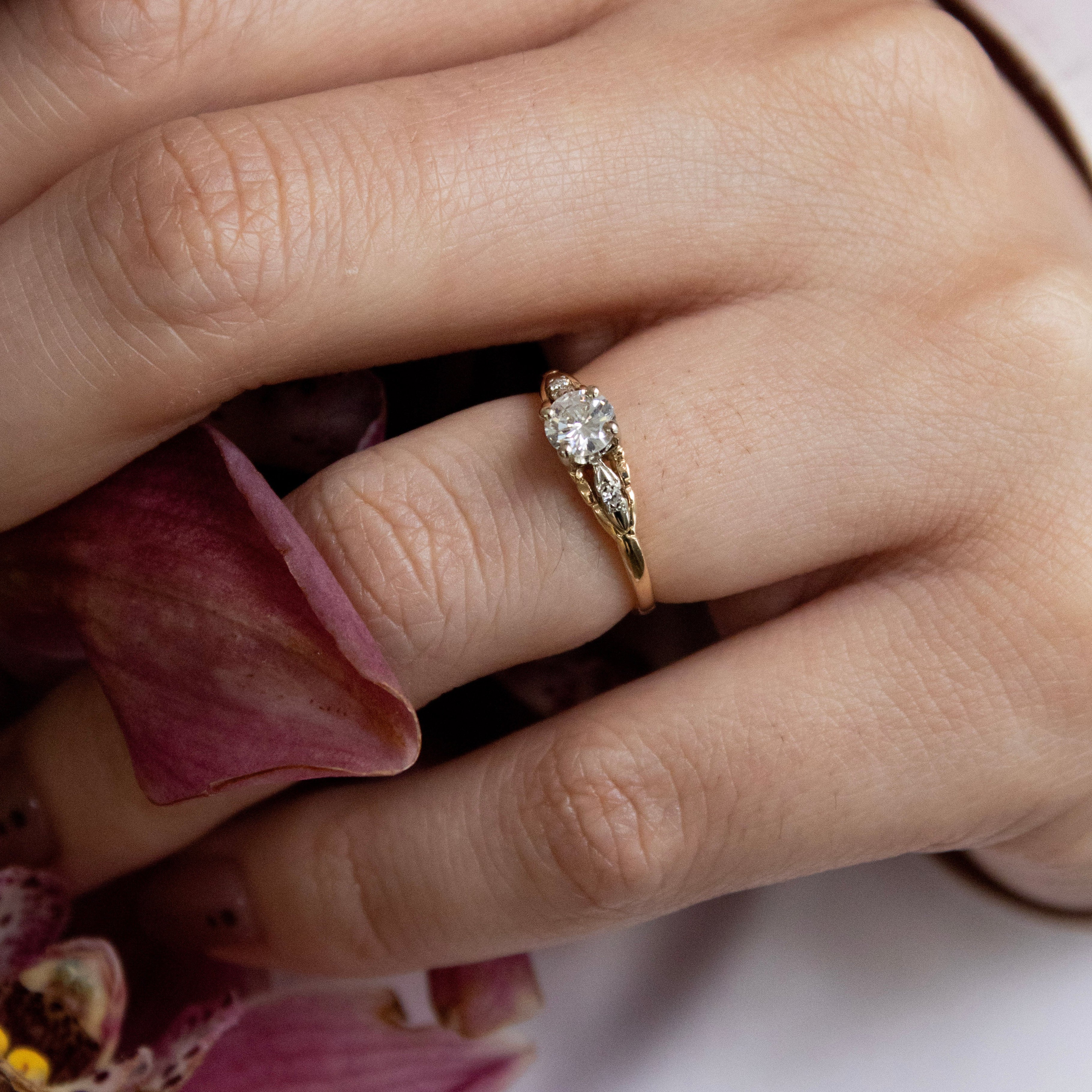 Close-up of a hand wearing a gold ring with a diamond, holding pink flowers.