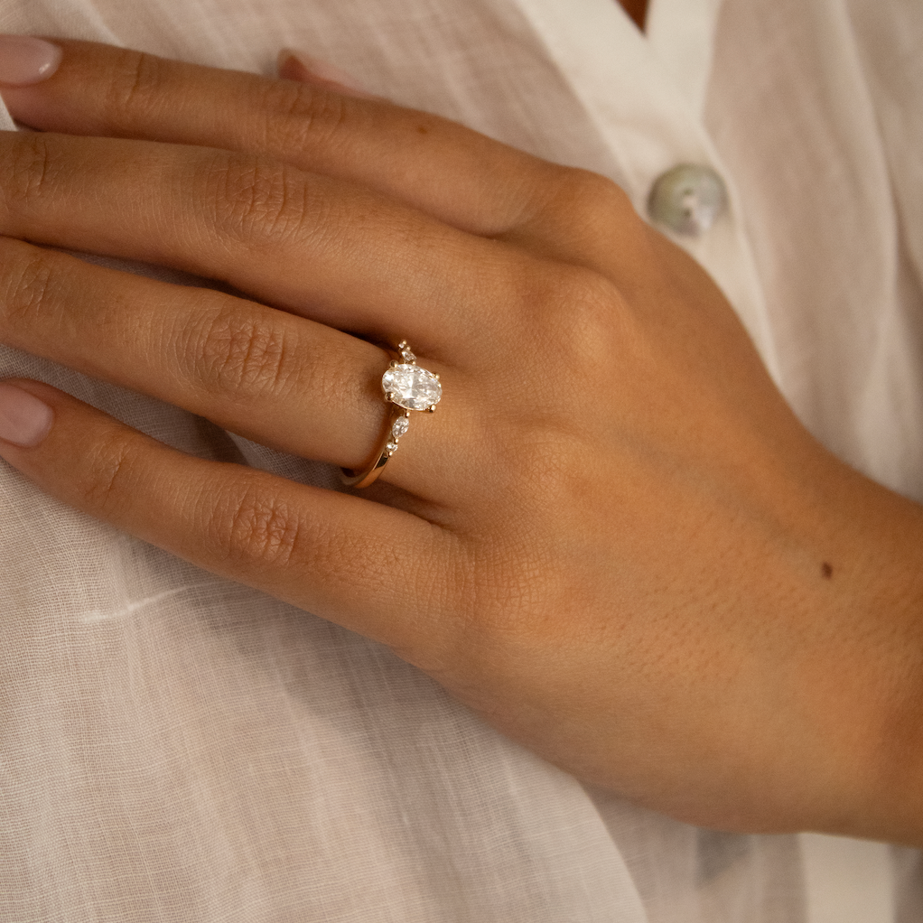 A close-up of a person's hand wearing a gold engagement ring with a marquise-cut center stone and small side stones, set against a white linen shirt.