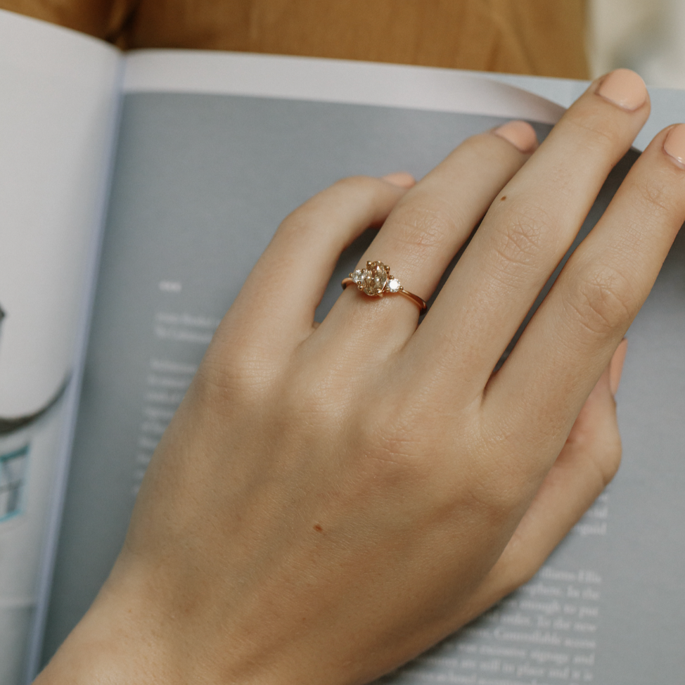 A close-up of a person's hand resting on an open book, wearing a yellow gold ring with diamond side stones.