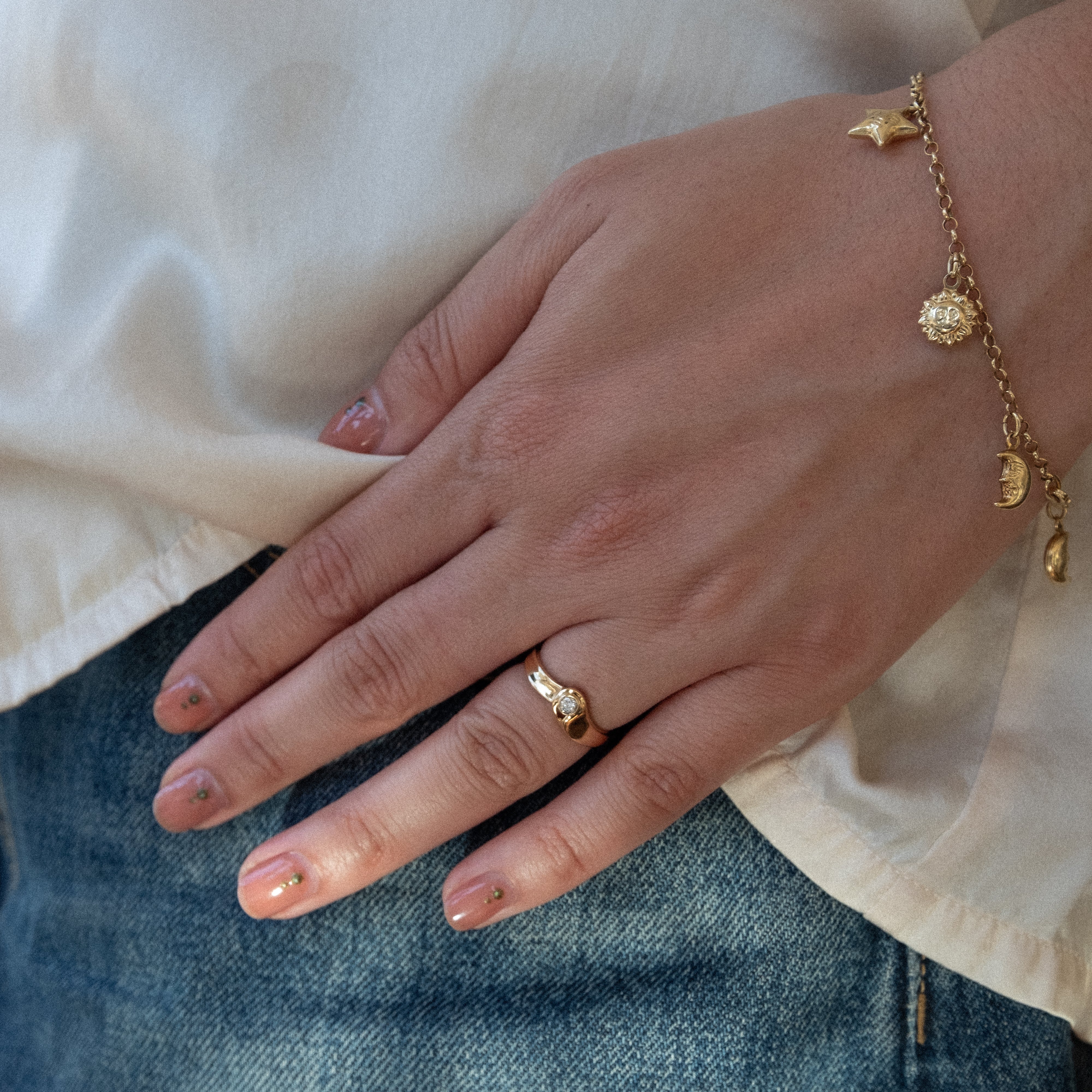 Hand wearing a gold ring and bracelet with a white shirt and blue jeans in the background