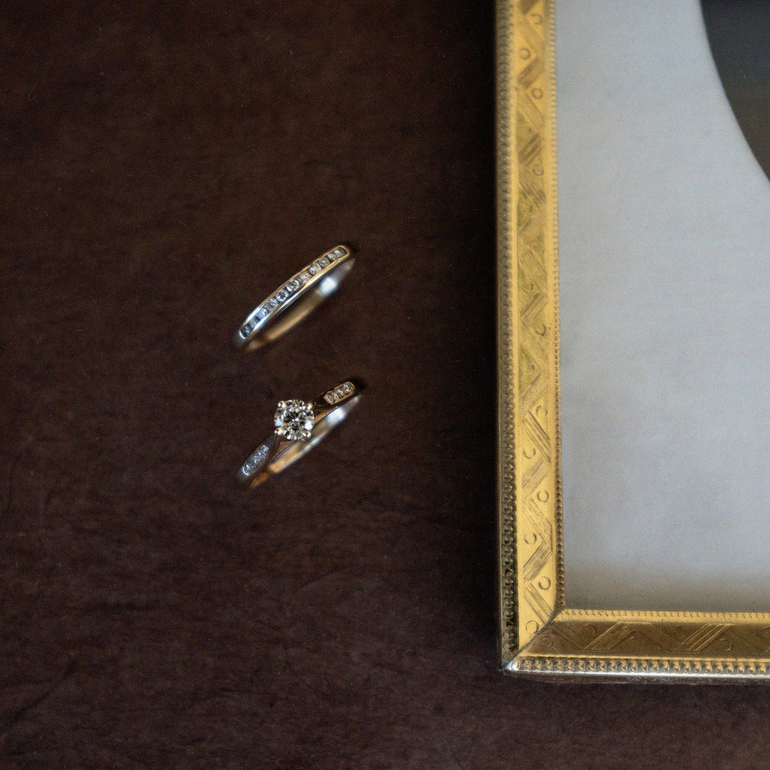 Two gold rings with diamonds on a dark surface next to an ornate gold frame.