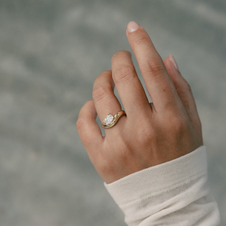 A close-up of a person's hand wearing a gold ring with a clear central gemstone, set against a soft, blurred gray background.