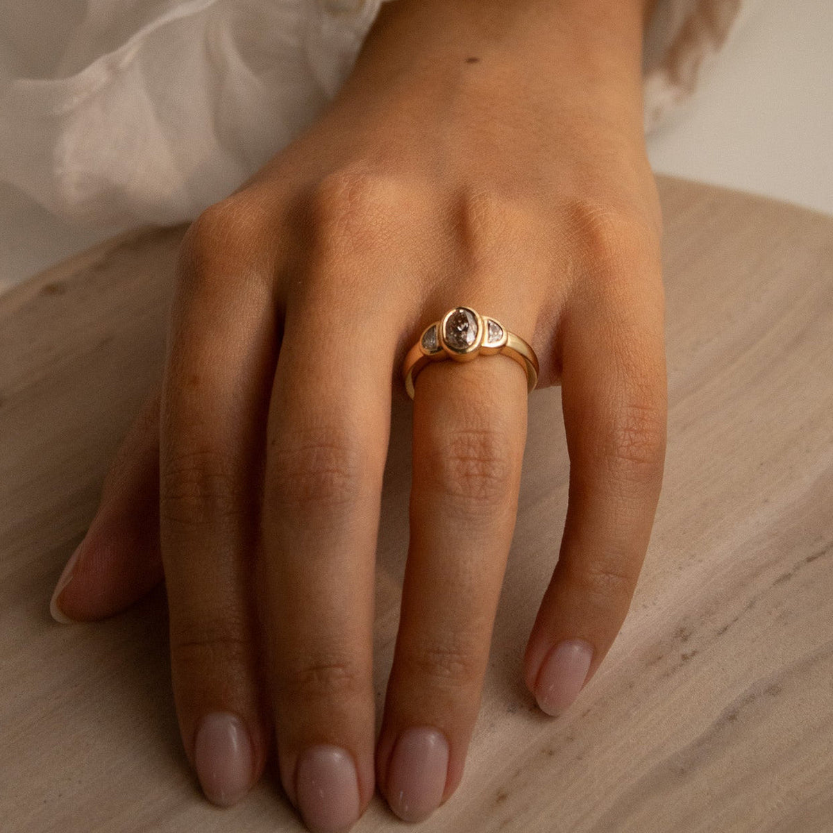 A gold ring with a round clear gemstone worn on a person's ring finger, shown with a white ruffled sleeve and a light wood surface in the background.